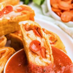 A sliced pizza roll with pepperoni and cheese, dipped in red marinara sauce, on a white plate. A bowl of pepperoni slices and fresh greens are blurred in the background.
