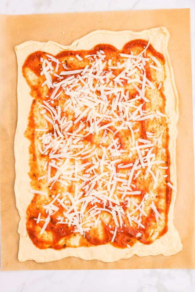 Rectangular pizza dough topped with tomato sauce and scattered shredded cheese, laid on a sheet of parchment paper.