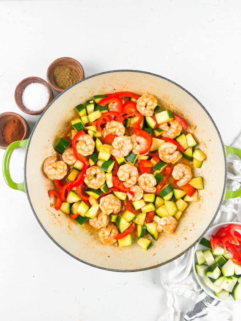 A large green-handled pot filled with shrimp, zucchini, and red bell peppers. Next to the pot are small bowls containing spices and salts. A small pile of chopped vegetables is on the side on a white surface.