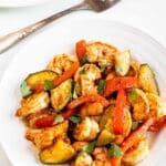 A white plate filled with cooked shrimp, sliced zucchini, and red bell peppers, garnished with herbs. Another partial plate and a fork are visible in the background, all set on a light surface.