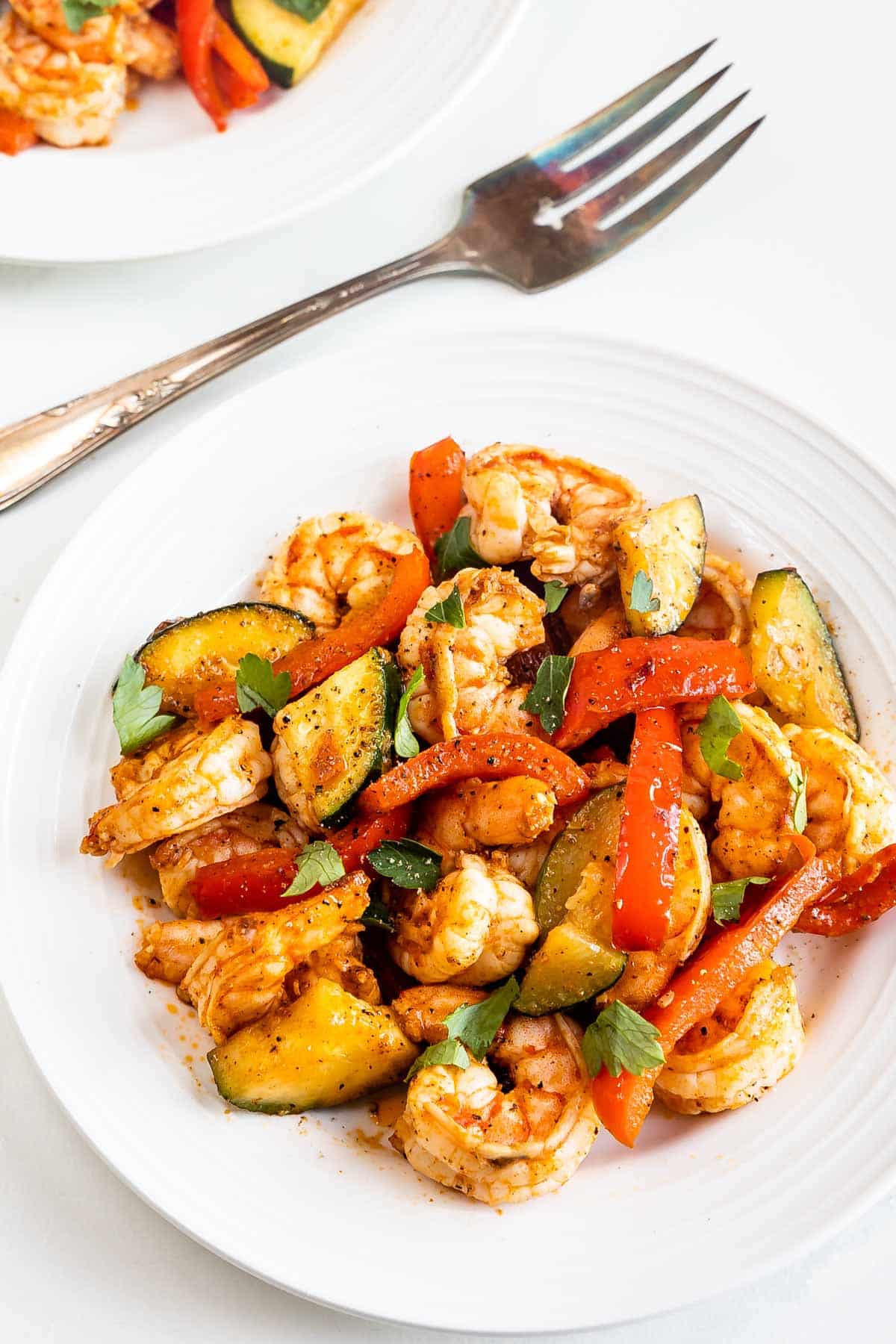 A white plate filled with cooked shrimp, sliced zucchini, and red bell peppers, garnished with herbs. Another partial plate and a fork are visible in the background, all set on a light surface.