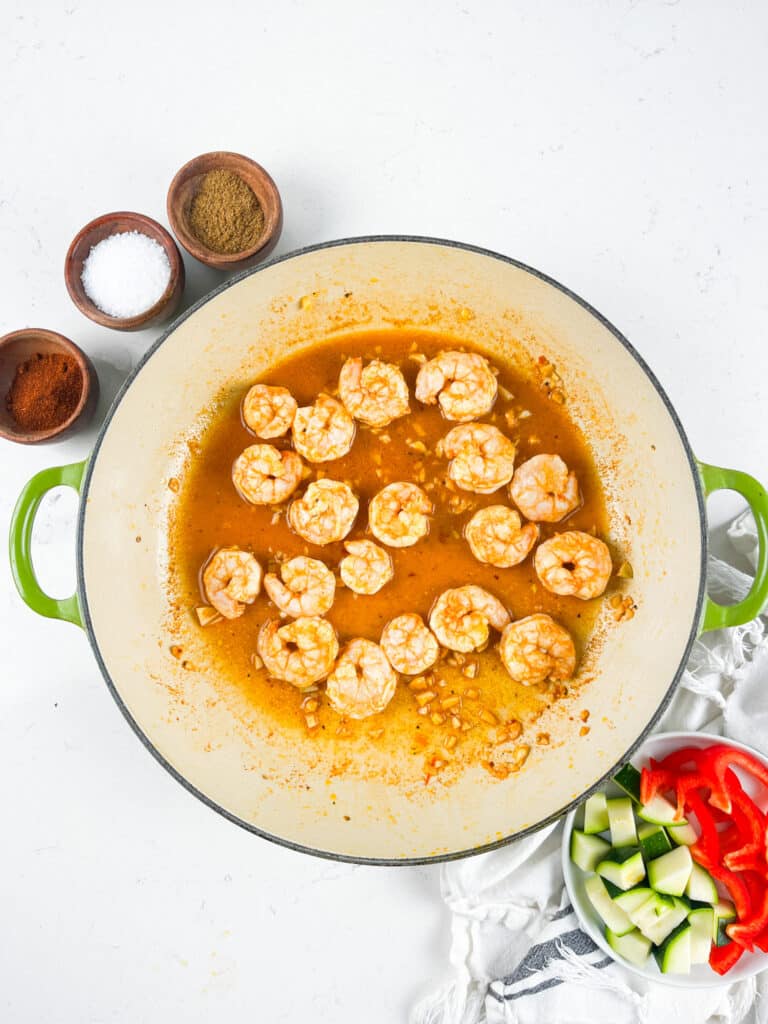 A pan with shrimp cooking in a brown sauce is centered on a white surface. Nearby are bowls of spices, sliced red peppers, and diced cucumbers. A striped cloth napkin is also visible.