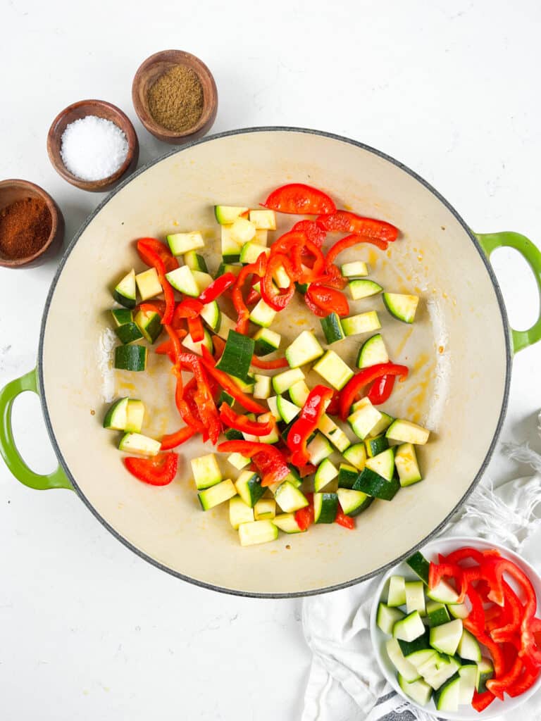 Chopped zucchini and red bell peppers cooking in a large, round green-handled pan. Nearby, small bowls hold spices and salt. A dishcloth and a small bowl of additional chopped zucchini rest on the white surface.