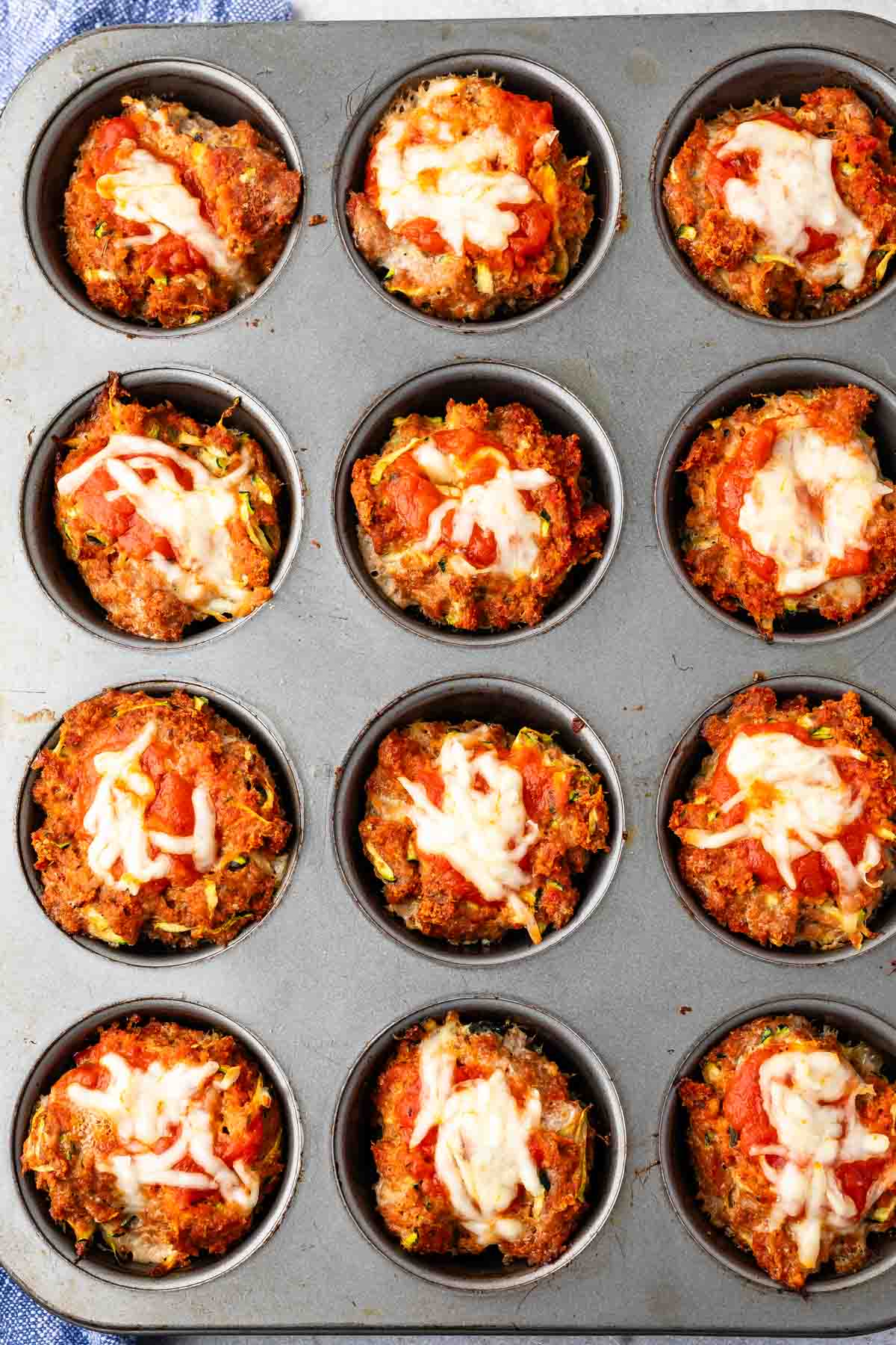 Twelve mini meatloaves in a muffin tin, each topped with melted cheese and tomato sauce. The meatloaves have a crispy, browned exterior and are evenly spaced in the round compartments of the tin.