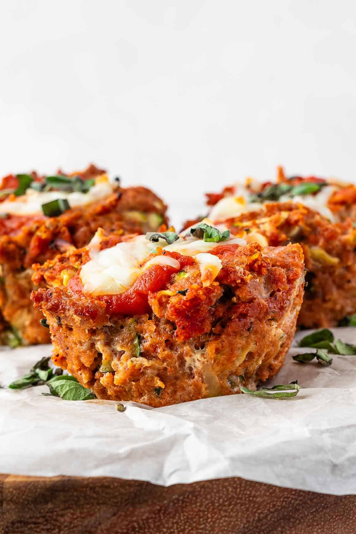 Four mini meatloaves topped with melted cheese and tomato sauce, garnished with fresh basil, displayed on parchment paper.