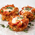 Three savory muffins topped with tomato sauce and melted cheese, garnished with chopped herbs, are arranged on crumpled parchment paper. The muffins have a golden-brown crust and visible vegetable pieces.