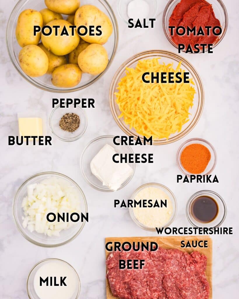 A variety of cooking ingredients are displayed on a white surface. Items include potatoes, salt, tomato paste, shredded cheese, pepper, butter, cream cheese, paprika, onion, milk, Parmesan, Worcestershire sauce, and ground beef.