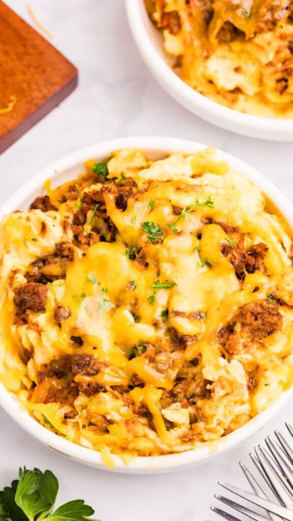 A bowl of cheesy mashed potatoes topped with ground beef and garnished with parsley. A fork rests in the foreground, and a second dish is partially visible in the background.
