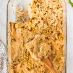 A glass baking dish holds a partially served chicken casserole with a golden crust, featuring chunks of chicken and cheese. A wooden spoon rests in the dish, surrounded by a patterned towel and sprigs of fresh parsley on a white marble surface.