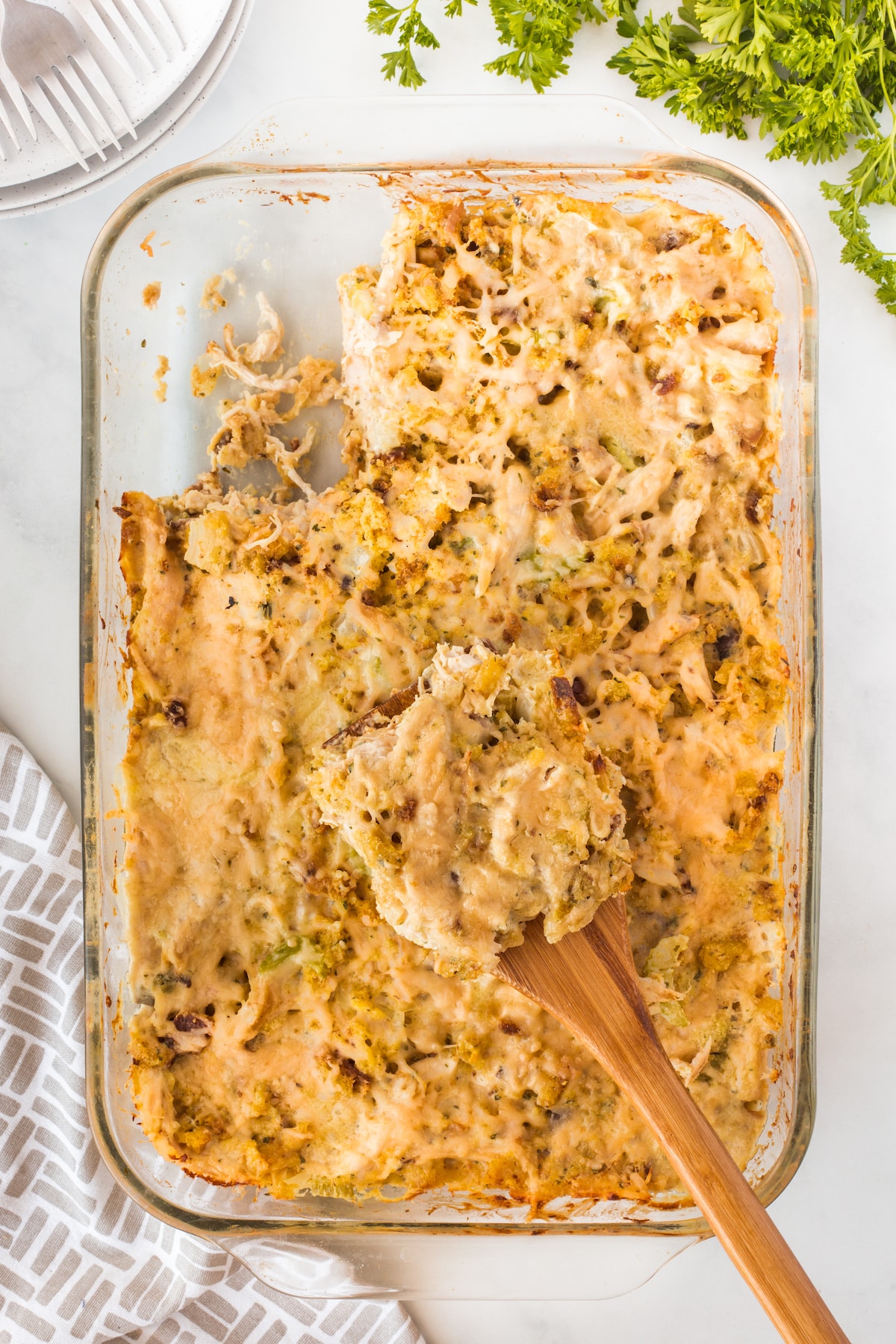 A glass baking dish holds a partially served chicken casserole with a golden crust, featuring chunks of chicken and cheese. A wooden spoon rests in the dish, surrounded by a patterned towel and sprigs of fresh parsley on a white marble surface.