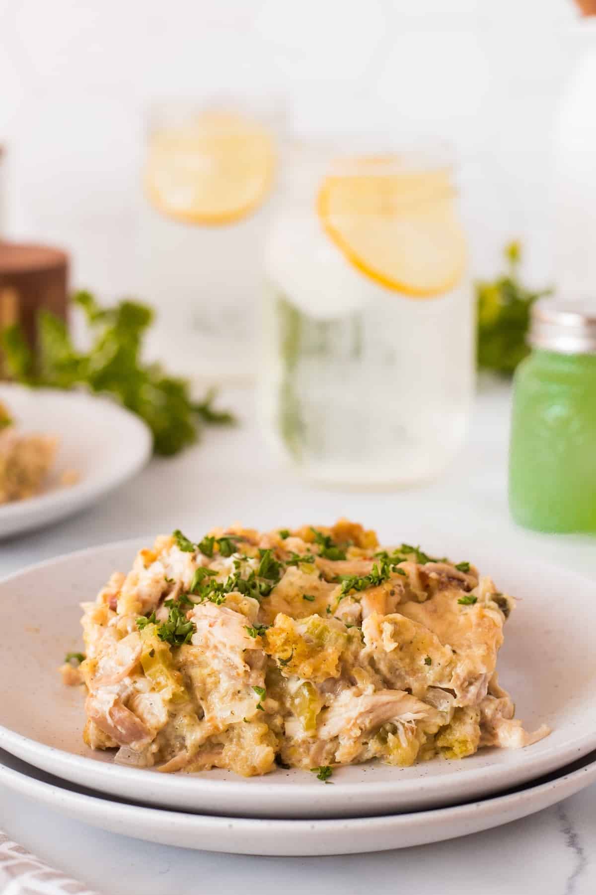 A plate of creamy chicken casserole garnished with chopped parsley is served on a white plate. In the background, there are glasses of lemon-infused water and a small green condiment container on a light-colored table.