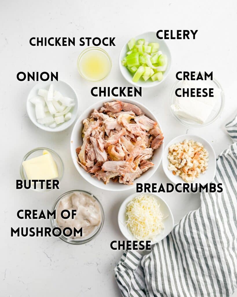 Ingredients arranged on a white surface with labels: chicken, celery, cream cheese, breadcrumbs, cheese, cream of mushroom, butter, onion, and chicken stock. A striped cloth is partially visible on the side.