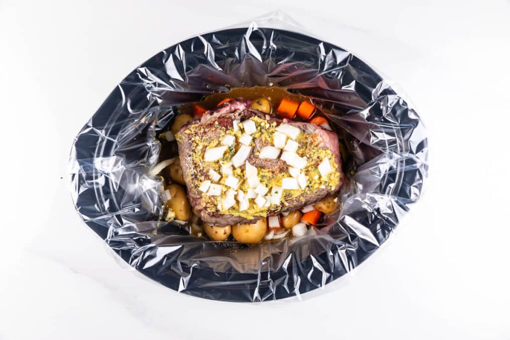 A slow cooker lined with a clear plastic liner, containing a seasoned pot roast, chopped white onions, baby potatoes, and sliced carrots. The ingredients are uncooked and neatly arranged, viewed from above.