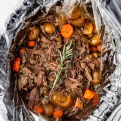 A slow cooker lined with a plastic bag contains a cooked pot roast. The dish includes shredded beef, potatoes, and carrots, garnished with a sprig of rosemary. The cooker is placed on a light surface with a striped cloth nearby.