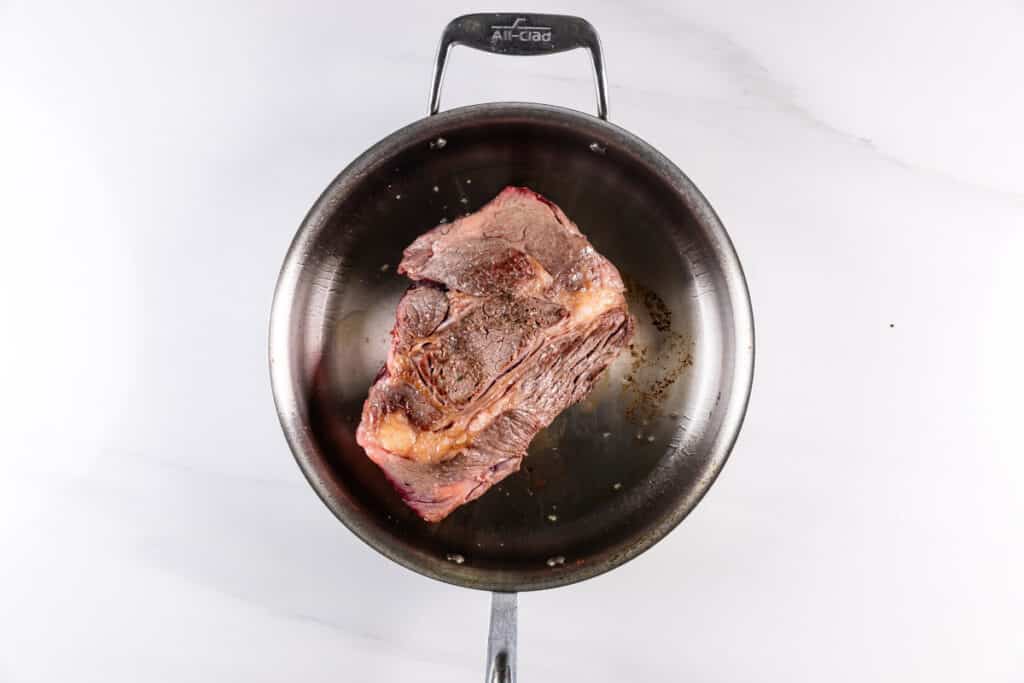 A large piece of seasoned beef searing in a round metal pan on a light countertop. The beef appears slightly browned, with visible marbling and a small amount of fat.