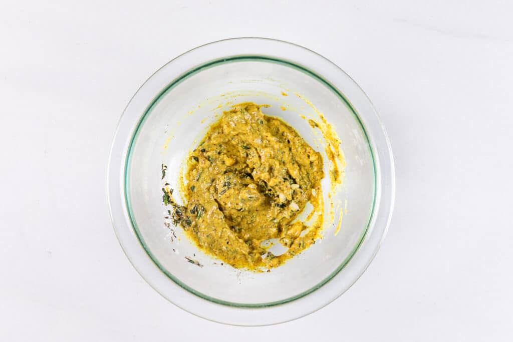 A glass bowl on a white surface contains a thick, yellow mixture with herbs or spices. The mixture appears partially blended, showcasing a textured surface.
