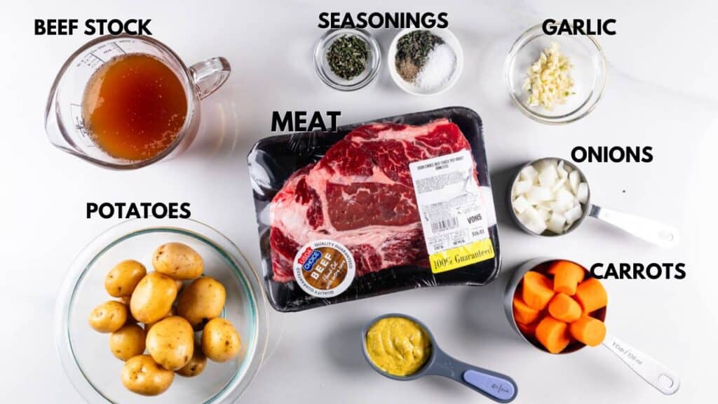 Top-down view of ingredients on a white surface: beef stock in a measuring cup, seasonings in small bowls, chopped garlic, diced onions, sliced carrots, a package of raw meat, whole potatoes in a bowl, and mustard in a measuring cup.