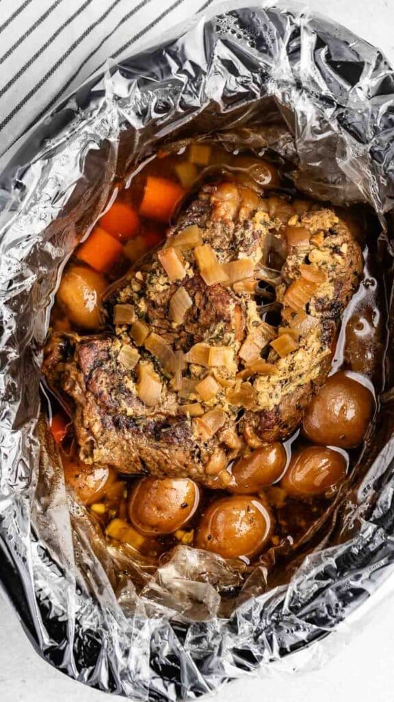 A roasted meat dish with browned meat, carrots, and potatoes is in a foil-lined slow cooker, surrounded by savory broth and topped with cooked onions. A striped cloth is partially visible in the background.