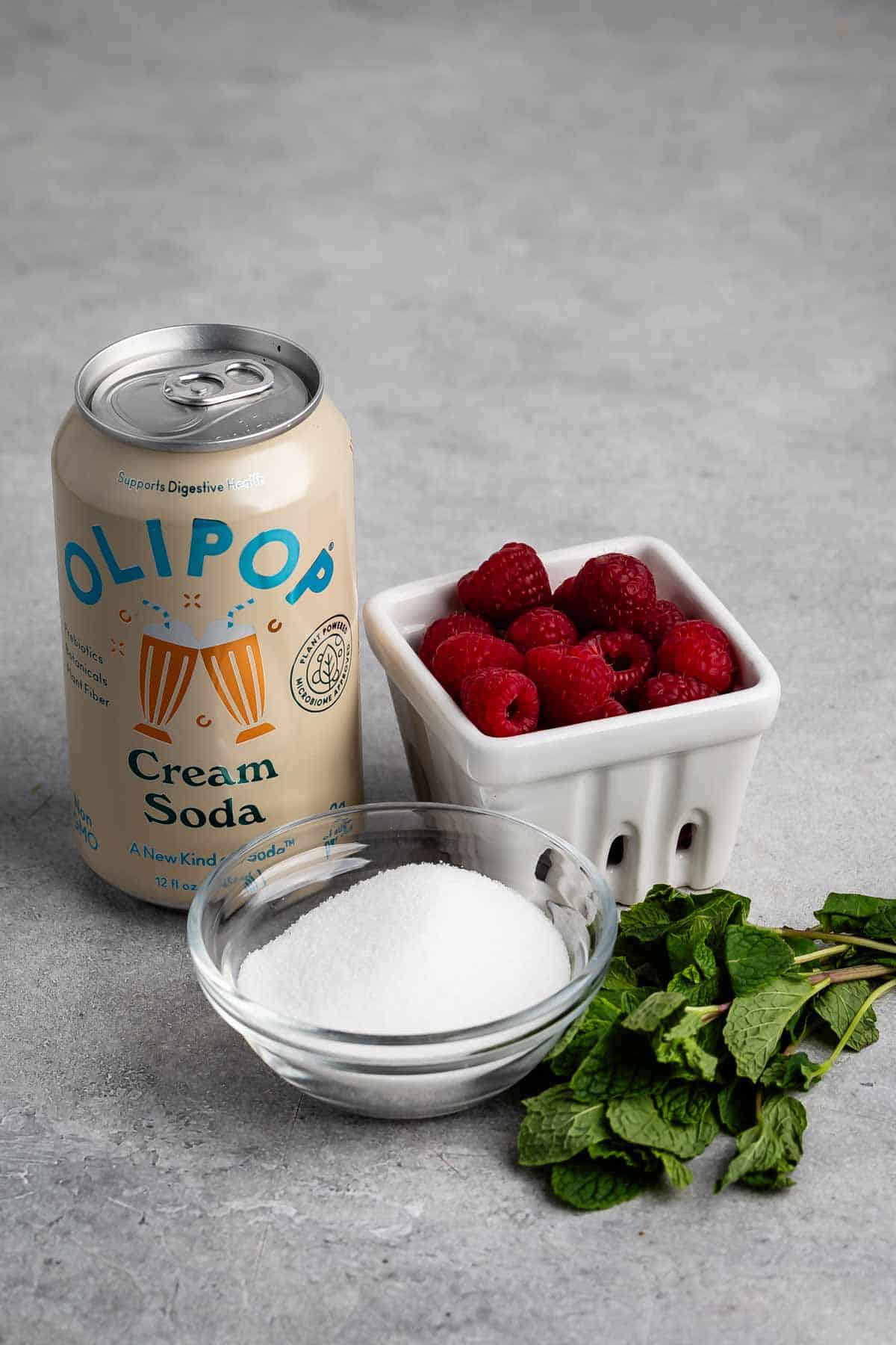 A can of Olipop cream soda is next to a ceramic container filled with fresh raspberries, a glass bowl containing white sugar, and a small bunch of mint leaves on a gray surface.