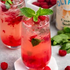 Two tall glasses of a pink raspberry cocktail with mint leaves on top. Fresh raspberries are scattered around, and a can of soda is in the background. The drink is garnished with mint and crushed ice.