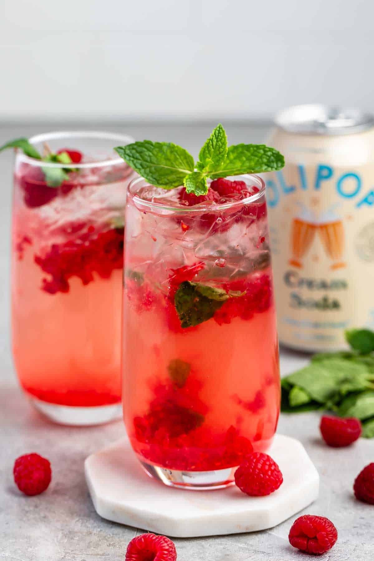 Two tall glasses of a pink raspberry mint drink are filled with ice and garnished with fresh mint and raspberries. A can of cream soda is in the background. Some fresh raspberries and mint leaves are scattered nearby on a light surface.