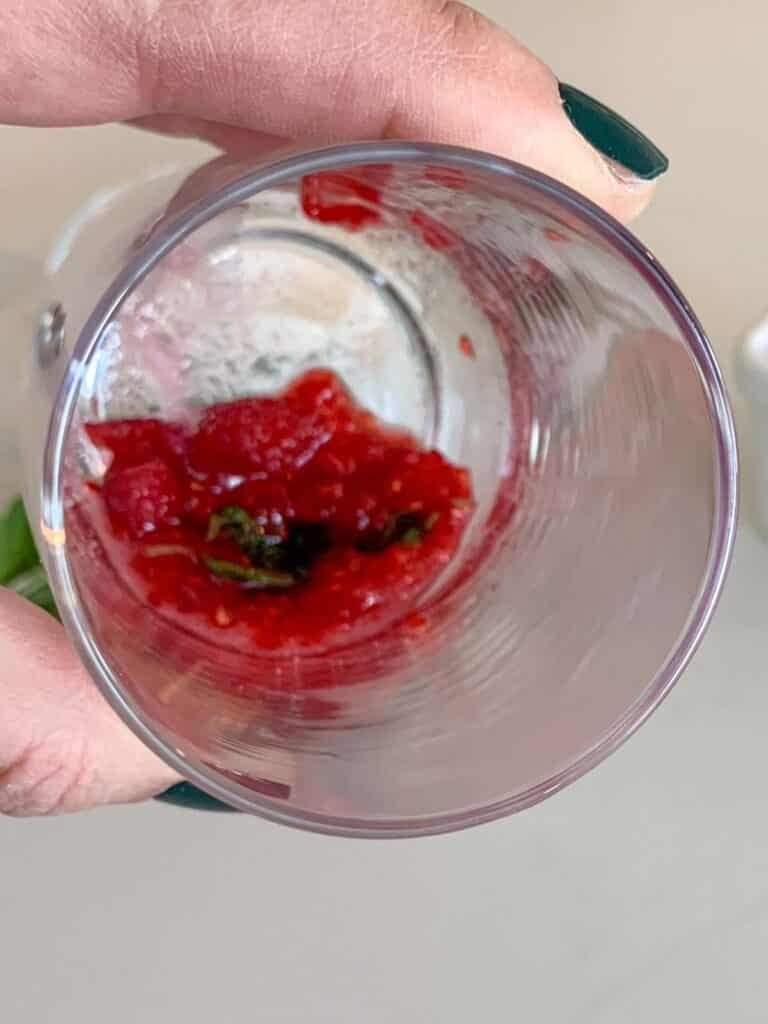 A hand with dark green nail polish holds a transparent glass from above, revealing red residue and a few leaves inside. The background is blurred, emphasizing the contents of the glass.