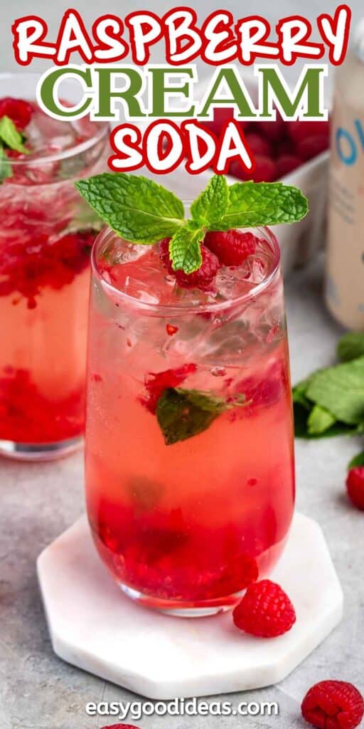 A refreshing glass of raspberry cream soda with ice, garnished with fresh raspberries and mint leaves. The pink drink sits on a white coaster, surrounded by a backdrop of additional raspberries and mint. Text reads RASPBERRY CREAM SODA.