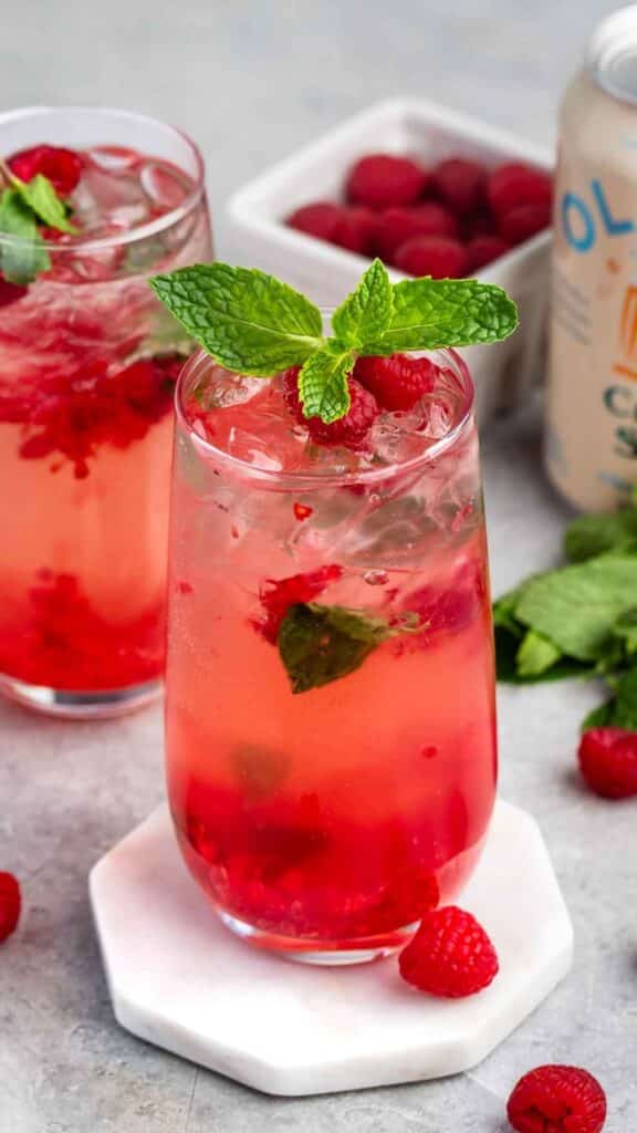 Two glasses of raspberry mojito garnished with mint leaves. The drinks are filled with crushed raspberries, ice, and mint. Fresh raspberries and a can are in the background, and a few raspberries are scattered on the surface.