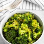 A bowl of seasoned broccoli florets sits on a gray surface. In the background, there is a striped cloth and a fork. The broccoli appears freshly cooked, with a vibrant green color and lightly toasted tips.