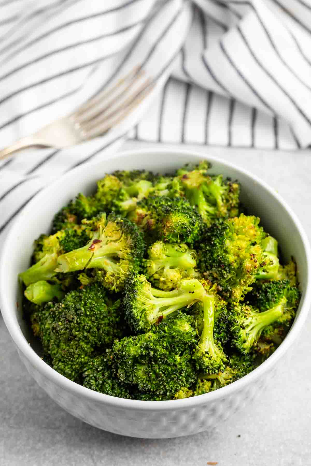 A bowl of seasoned broccoli florets sits on a gray surface. In the background, there is a striped cloth and a fork. The broccoli appears freshly cooked, with a vibrant green color and lightly toasted tips.