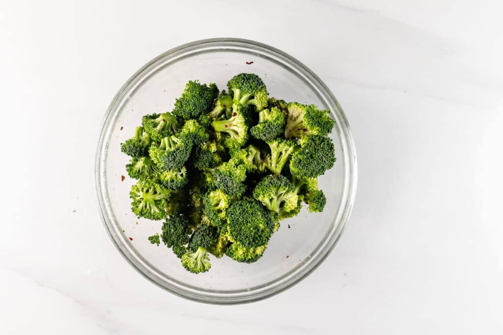 A clear glass bowl filled with fresh green broccoli florets on a white marble surface.