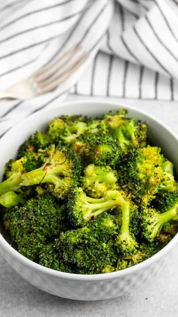 A bowl of seasoned roasted broccoli florets is placed on a light gray surface with a white and black striped cloth and a fork in the background.