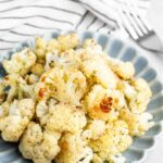 Roasted cauliflower florets seasoned with herbs and spices are served on a scalloped blue plate. A striped cloth and a fork rest in the background.