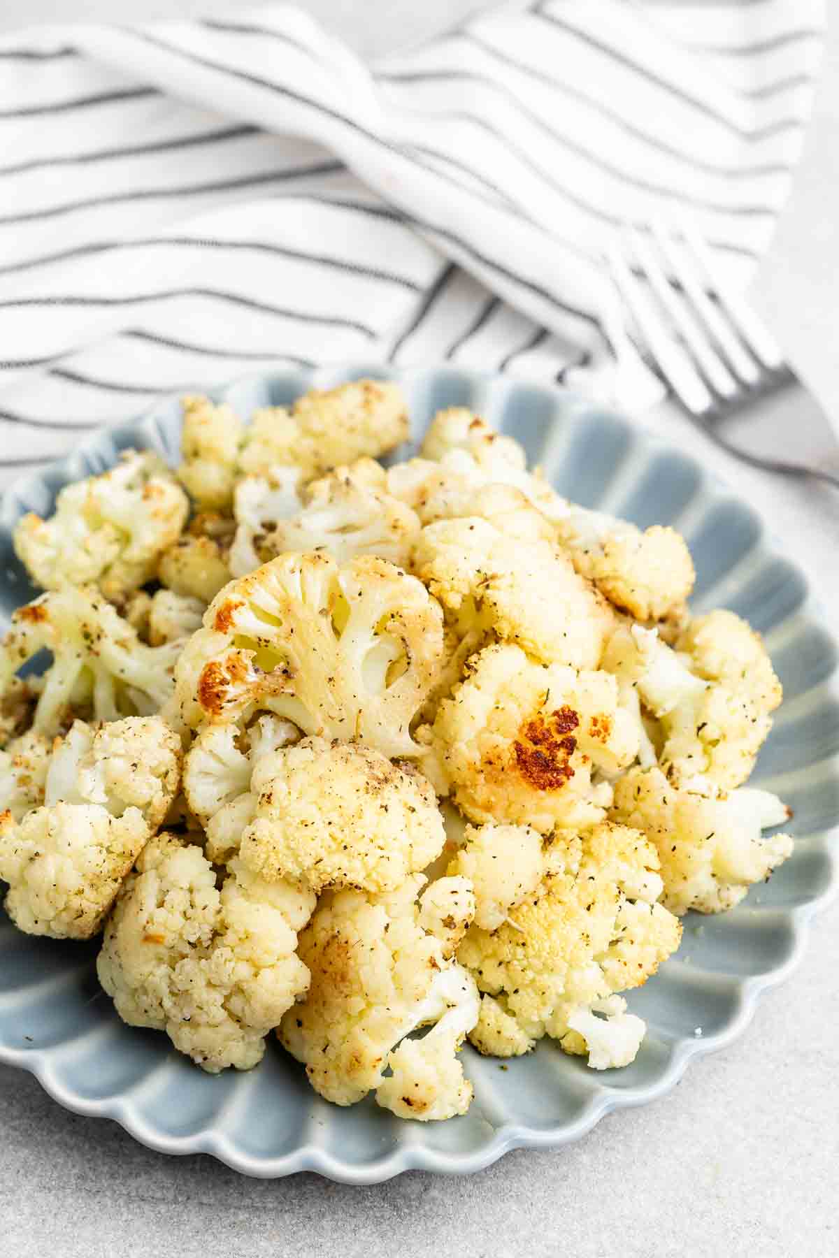 Roasted cauliflower florets seasoned with herbs and spices are served on a scalloped blue plate. A striped cloth and a fork rest in the background.