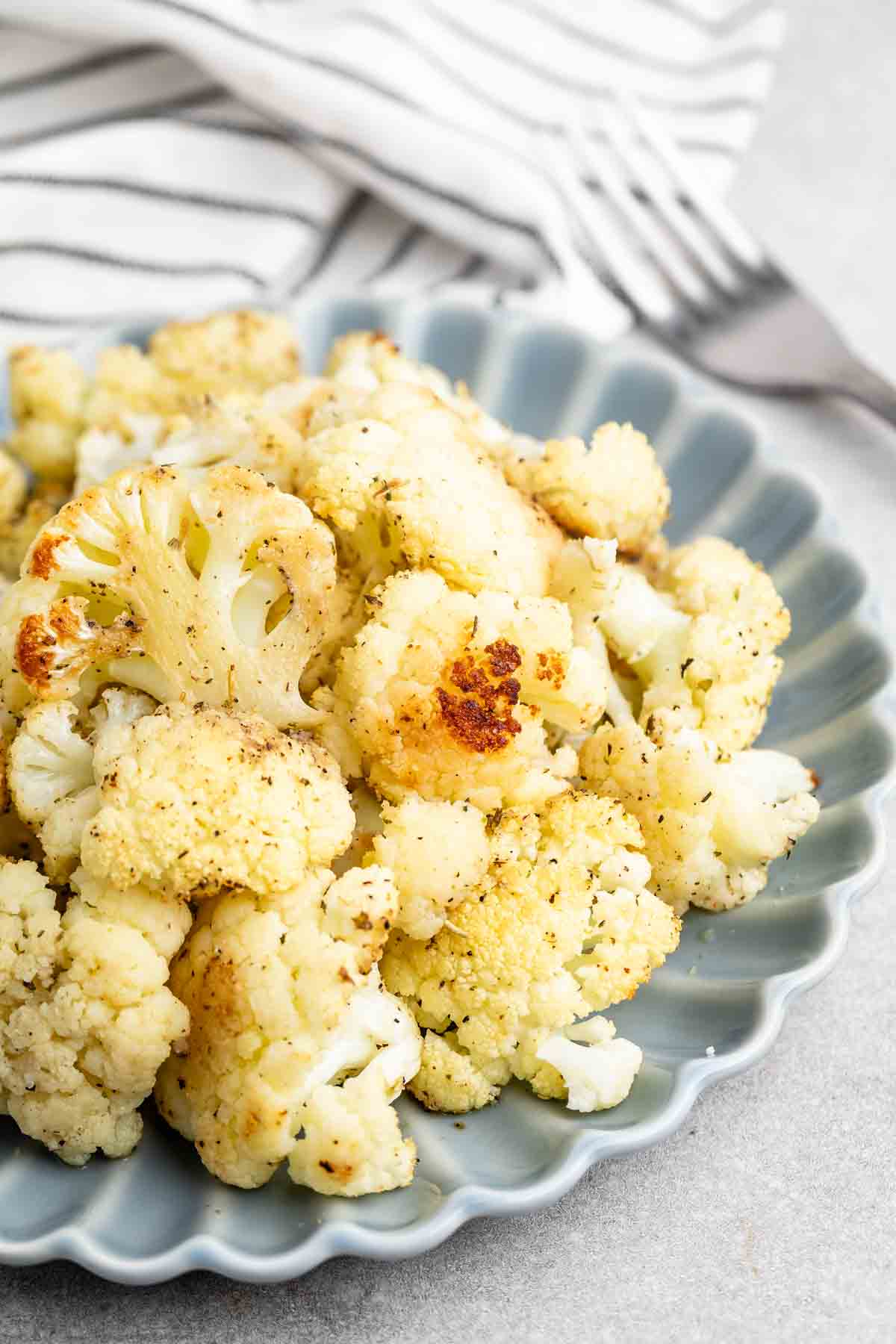 A plate filled with roasted cauliflower florets, lightly seasoned and browned, is displayed. A striped cloth and a fork are partially visible in the background.