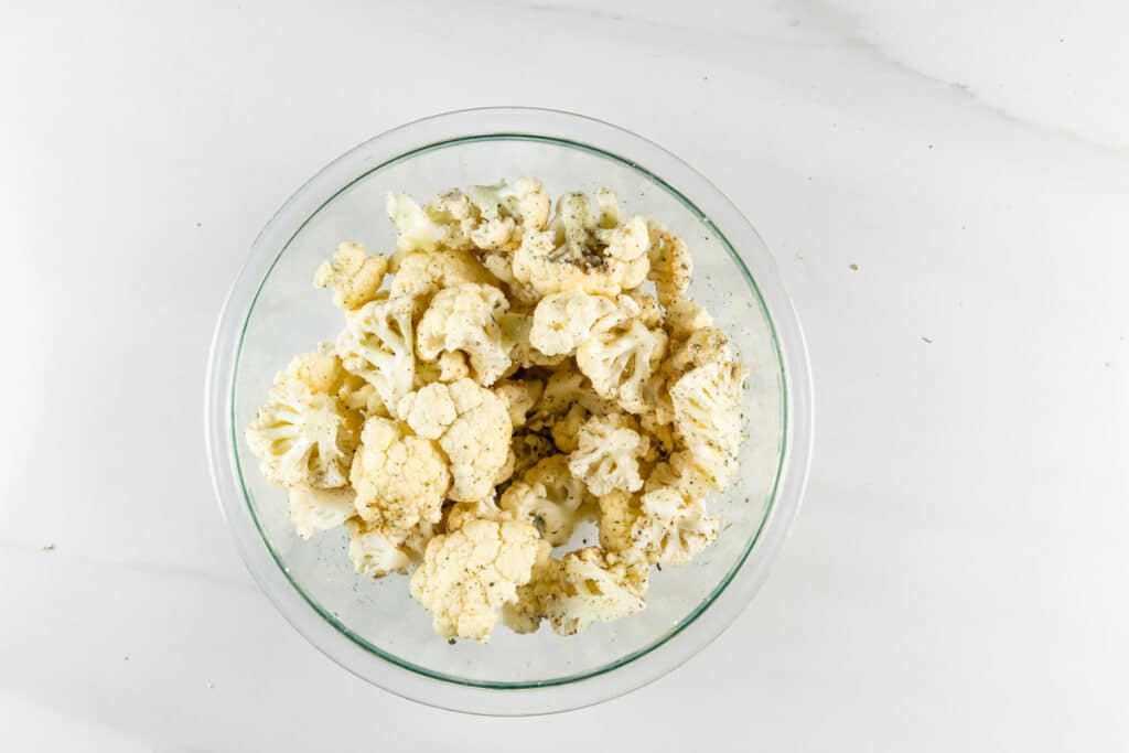 A glass bowl filled with cauliflower florets sprinkled with black pepper sits on a white marble surface.