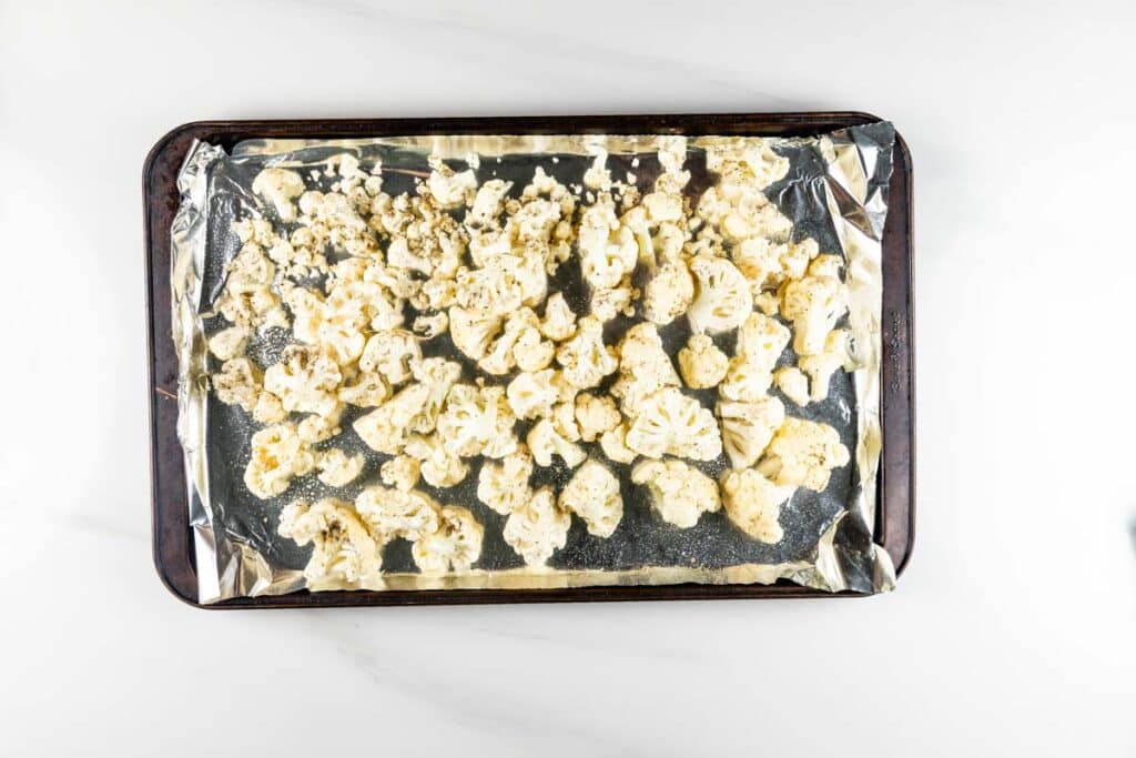 Chopped cauliflower florets spread on a foil-lined baking sheet, ready for roasting. The background is a light, neutral surface.