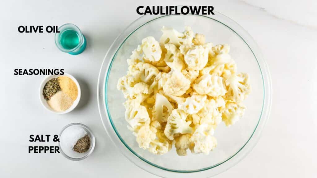 A bowl of cauliflower florets on a white surface. To the left, small bowls contain olive oil, mixed seasonings, and salt and pepper. Labels identify each ingredient.