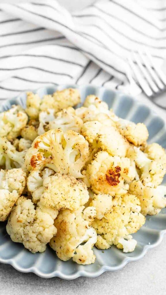 A plate of roasted cauliflower florets seasoned with pepper, placed on a gray scalloped dish. A white and black striped cloth with a fork lies in the background.