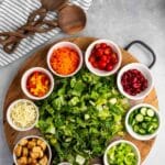 A round wooden board holds bowls of cherry tomatoes, shredded carrots, kidney beans, sliced cucumbers, celery, nuts, seeds, croutons, shredded cheese, diced bell peppers, and a pile of chopped lettuce. Two wooden utensils and a striped towel are nearby.