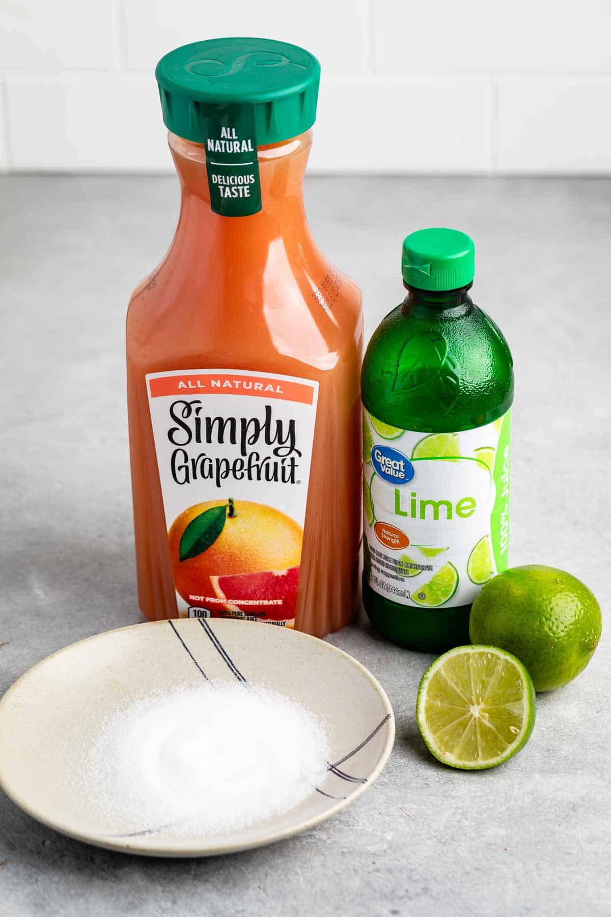 A bottle of Simply Grapefruit juice and a bottle of lime juice are on a countertop. Beside them are two whole limes, one cut in half, and a bowl containing sugar.