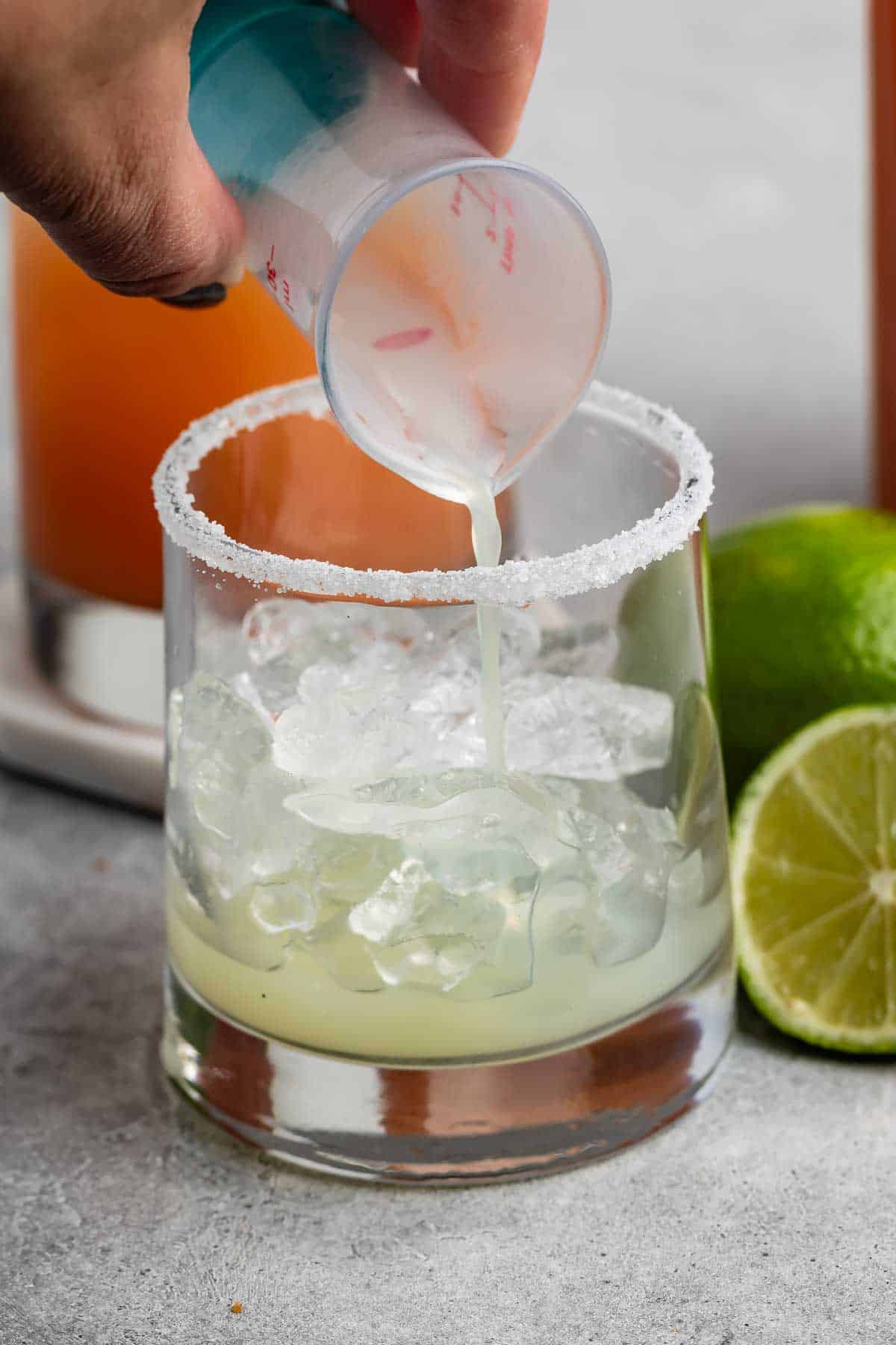 A hand pours a creamy liquid from a small cup into a glass with ice cubes. The glass has a sugar rim, and there are fresh lime halves nearby. A blurred drink in the background hints at a cocktail-making scene.