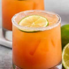 A close-up of a glass filled with an orange-colored cocktail, garnished with a lime slice on top and a sugared rim. Theres another similar glass in the background, with fresh lime halves beside them on a gray surface.