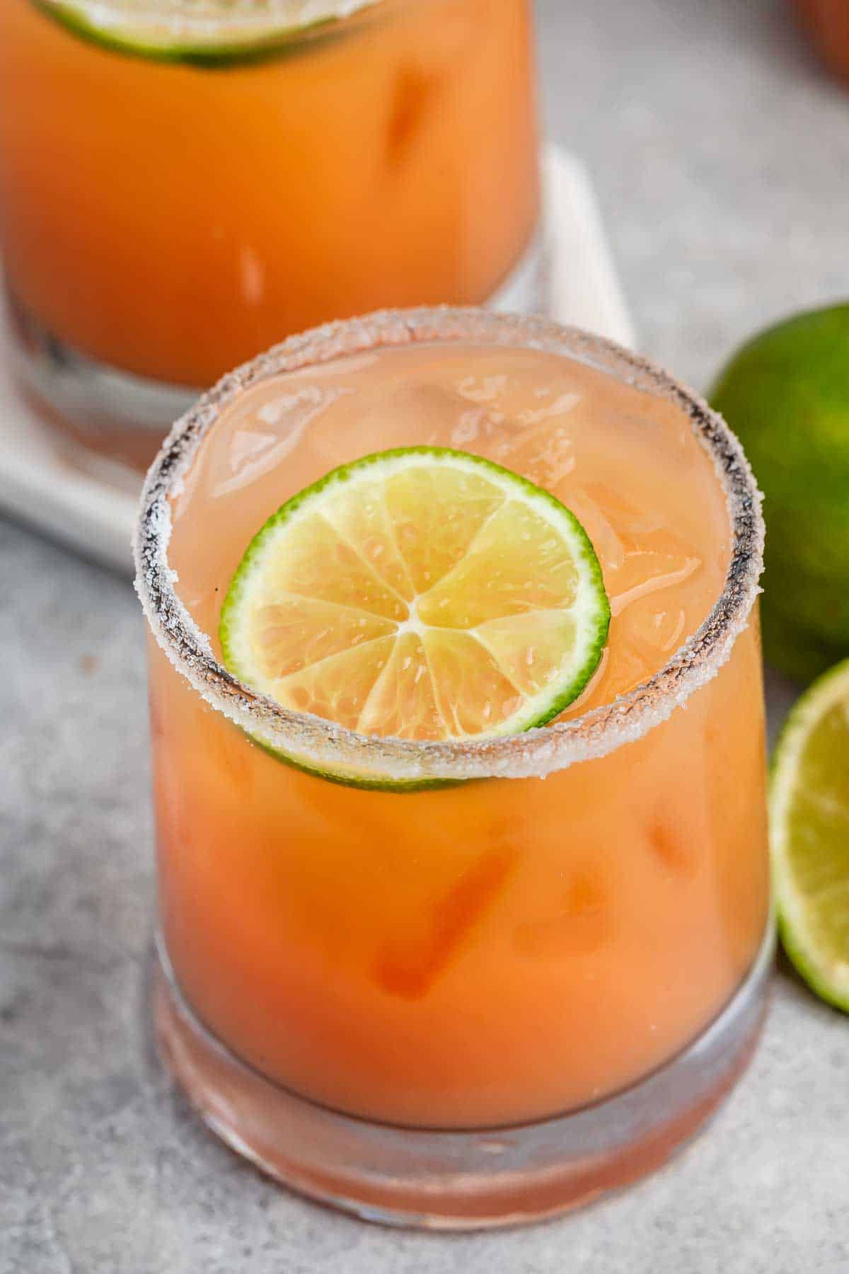 A glass filled with orange-hued liquid and ice, garnished with a slice of lime on the rim. The rim is coated with a fine, powdery substance, and a whole lime and a lime half are placed beside the glass.