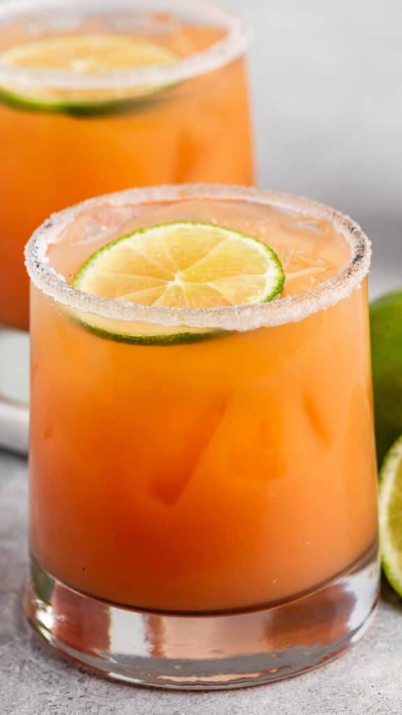 A close-up of a glass filled with an orange-colored cocktail, featuring a lime wheel on top. The rim of the glass is coated with sugar. Another similar drink is blurred in the background, alongside fresh lime halves on a gray surface.