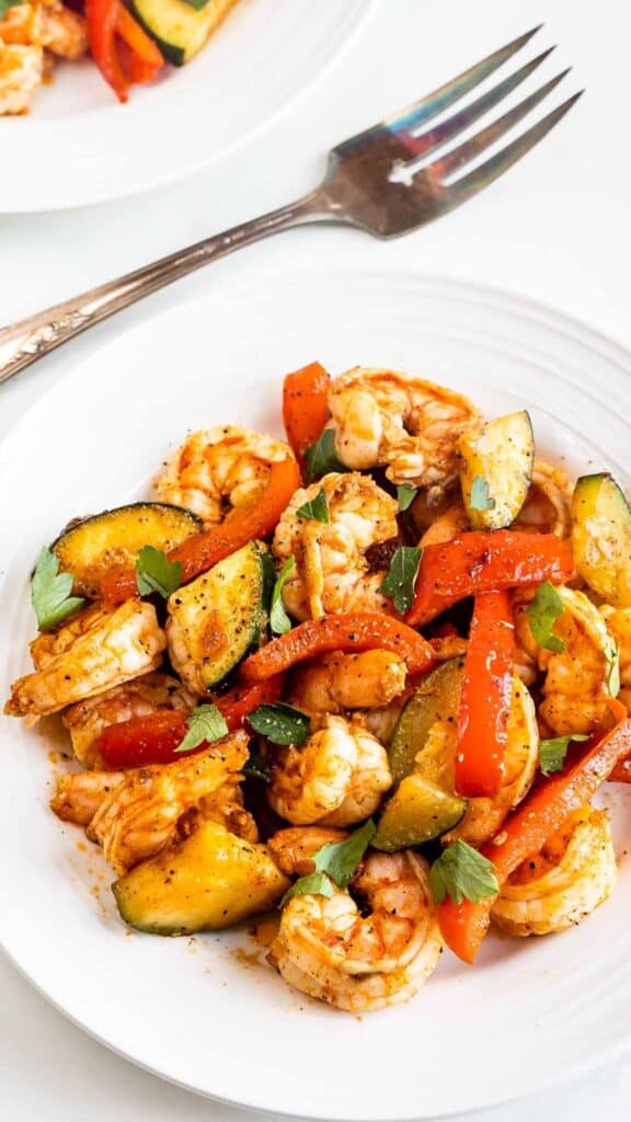 A white plate filled with shrimp, red bell peppers, and zucchini, garnished with fresh herbs. A fork rests beside the plate on a white surface.