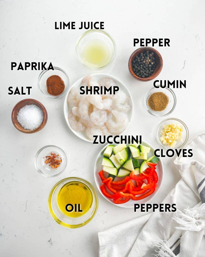 Top view of ingredients for a recipe labeled on a white surface. Includes olive oil, red pepper flakes, paprika, salt, lime juice, shrimp, diced zucchini, sliced red peppers, cumin, black pepper, minced garlic cloves. A white towel is partially visible.