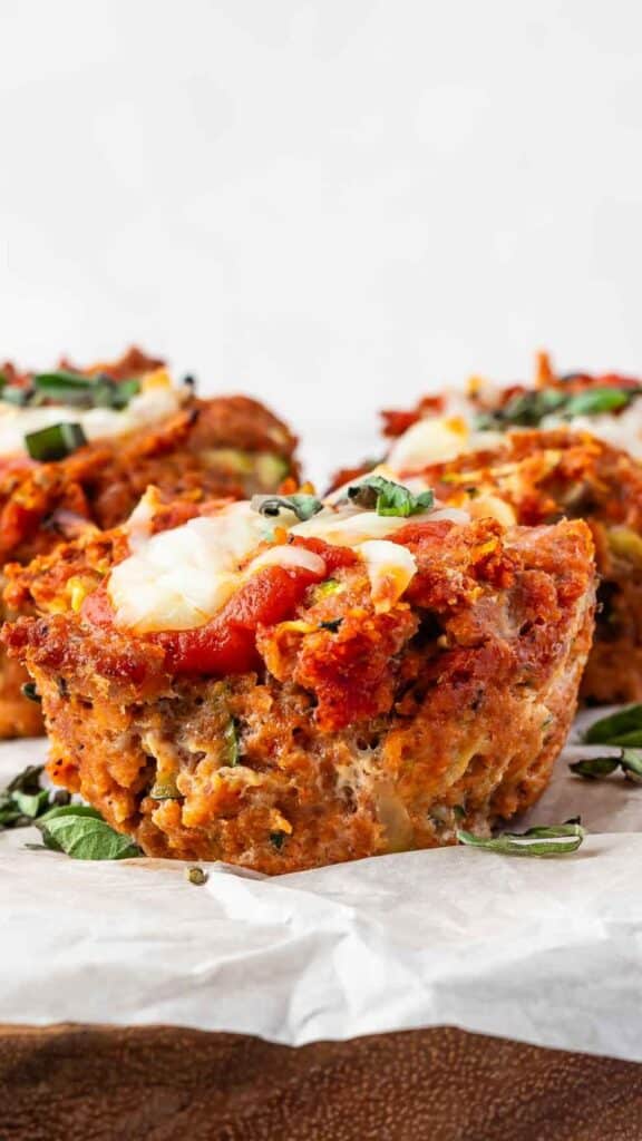 Close-up of individual meatloaves topped with tomato sauce and melted cheese, garnished with chopped herbs. They are placed on a white parchment paper, highlighting their crispy, golden-brown exterior and savory toppings.