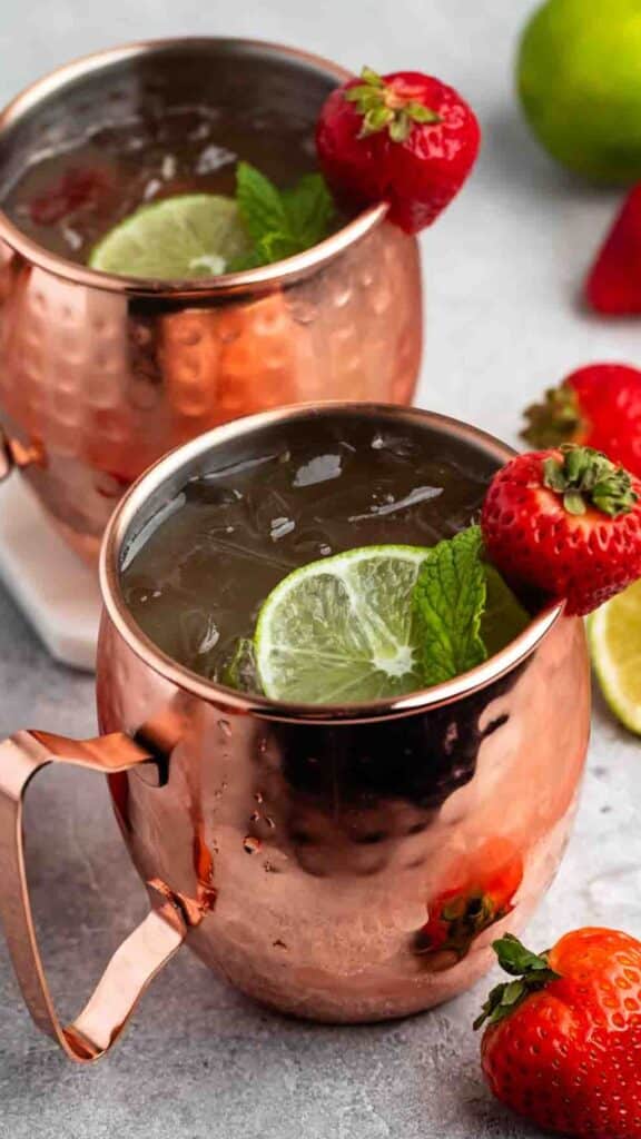 Two copper mugs filled with a refreshing beverage, garnished with lime slices, mint leaves, and fresh strawberries. The mugs are placed on a light surface with a lime and strawberries in the background.