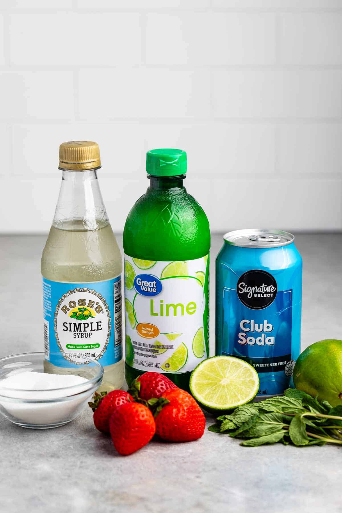 Bottles of simple syrup and lime juice, a can of club soda, a bowl of sugar, with fresh strawberries, a cut lime, a whole lime, and mint leaves on a light surface.